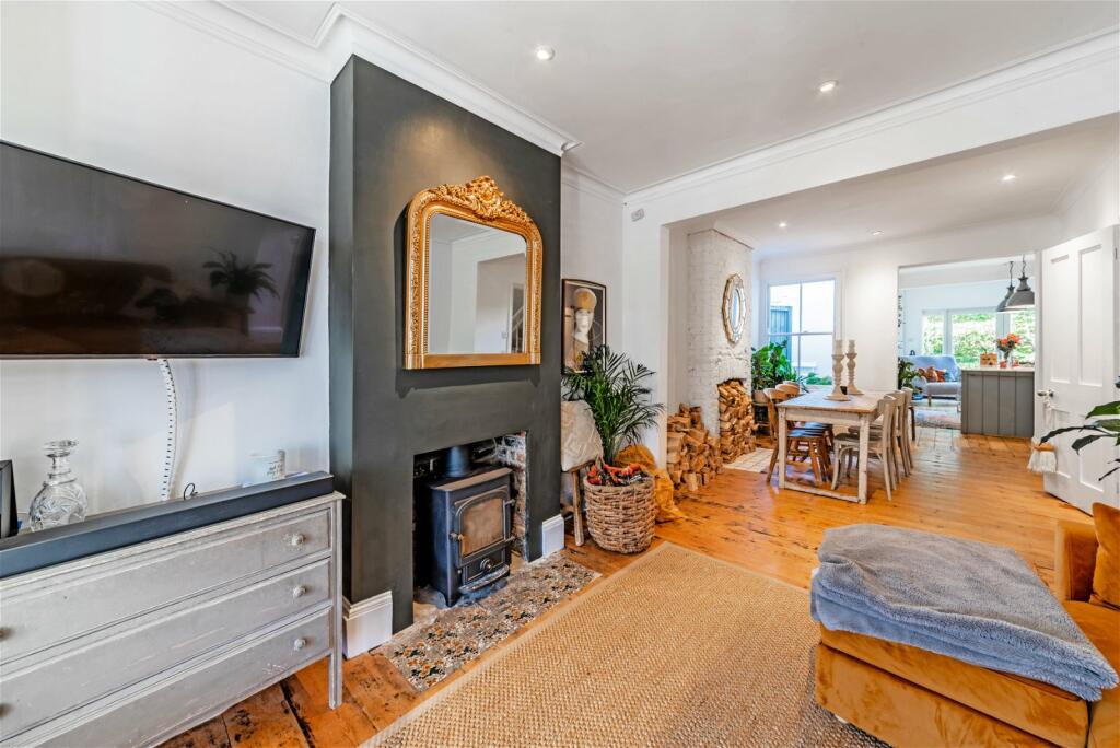 The living area is characterized by a dark accent wall featuring an ornate mirror above a traditional fireplace. A television is mounted on a light-colored cabinet. Natural wood flooring complements a cozy area rug and a plush throw is casually draped over a seat.