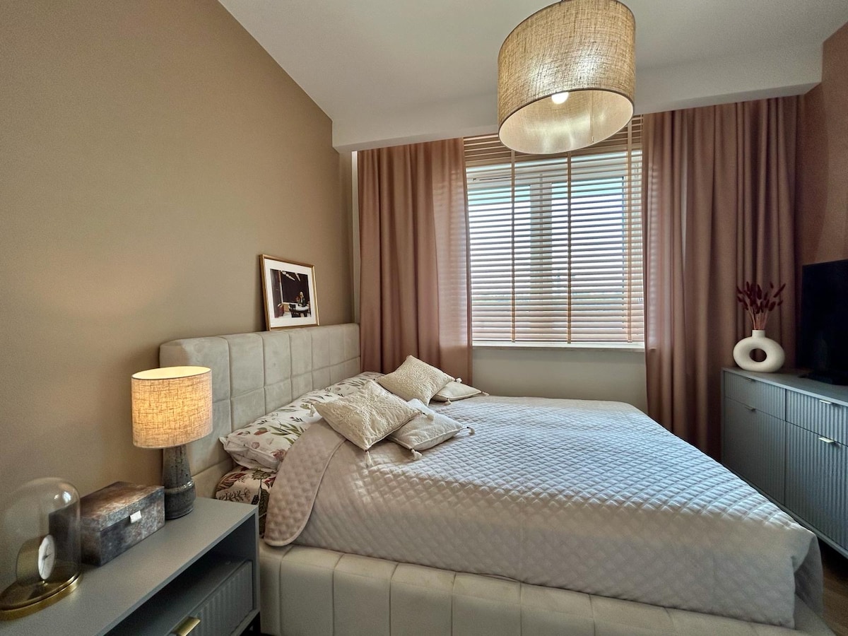 A serene bedroom features a spacious bed with light bedding, complemented by soft pillows. Natural light filters through slatted blinds, enhancing the calm atmosphere. A bedside table holds a lamp, and a textured light fixture hangs above. A dresser is positioned against the wall, providing additional storage.