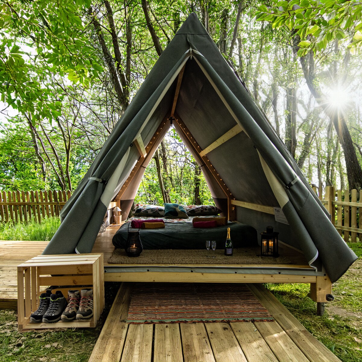 A tent with a triangular shape is set amidst trees, highlighted by natural light filtering through the foliage. The spacious interior features a comfortable bed adorned with cushions and soft blankets. An outdoor wooden deck with a small table enhances the inviting atmosphere.