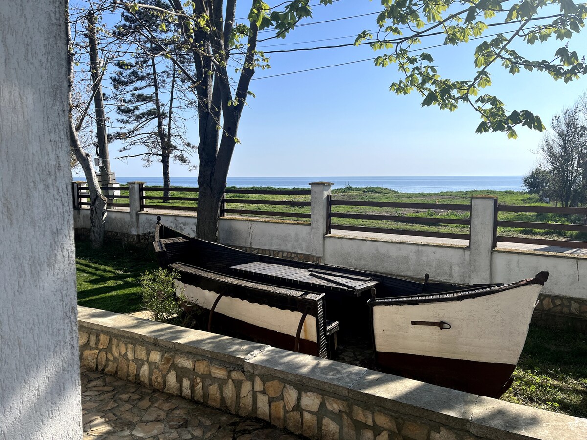 A traditional wooden boat is positioned in the garden, with a serene view of the sea visible in the background. The stone wall and grassy area provide a tranquil contrast, while trees frame the scene under clear blue skies.