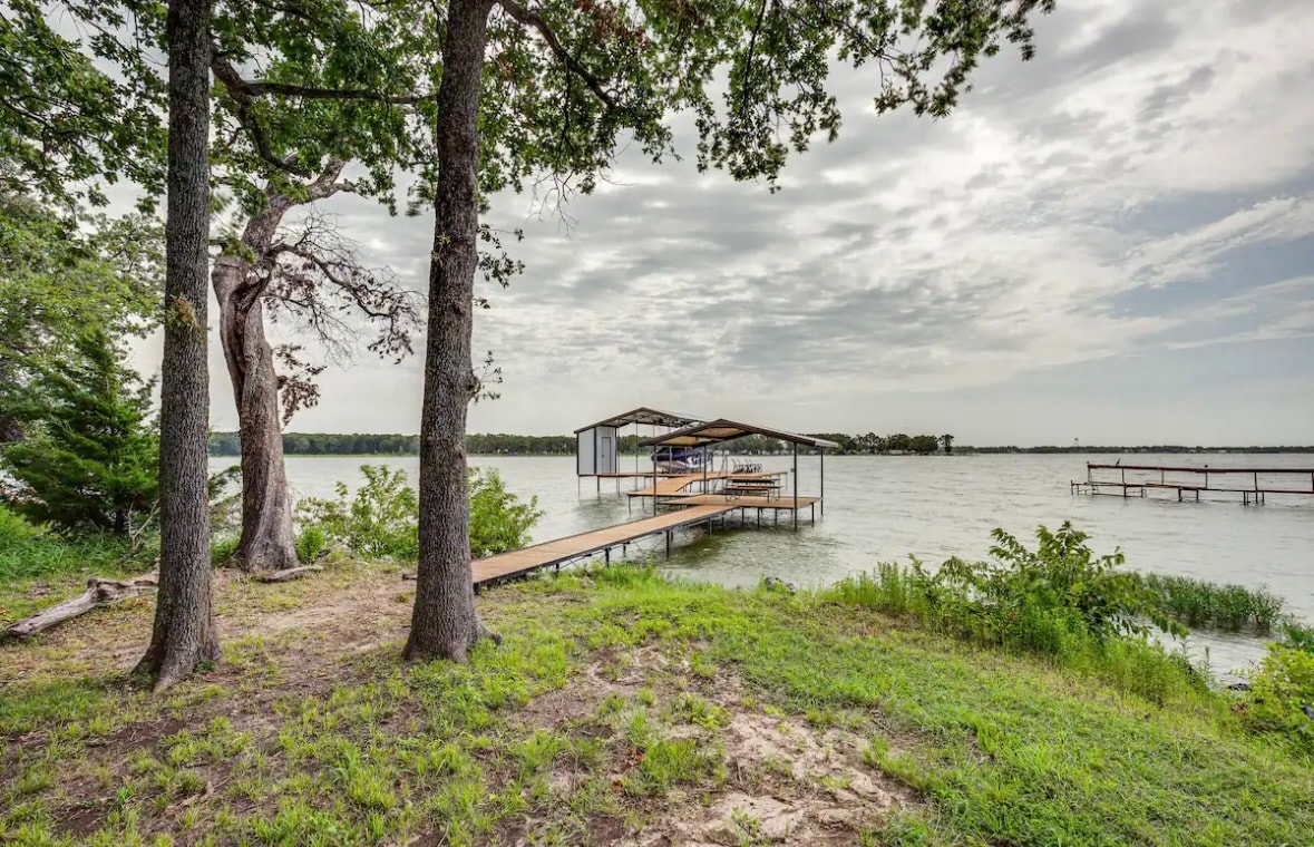 A serene lakeside view features a private dock extending over calm waters, surrounded by lush greenery and trees. The sky is partially cloudy, creating a peaceful atmosphere for outdoor activities. The dock serves as a perfect access point for swimming and fishing.