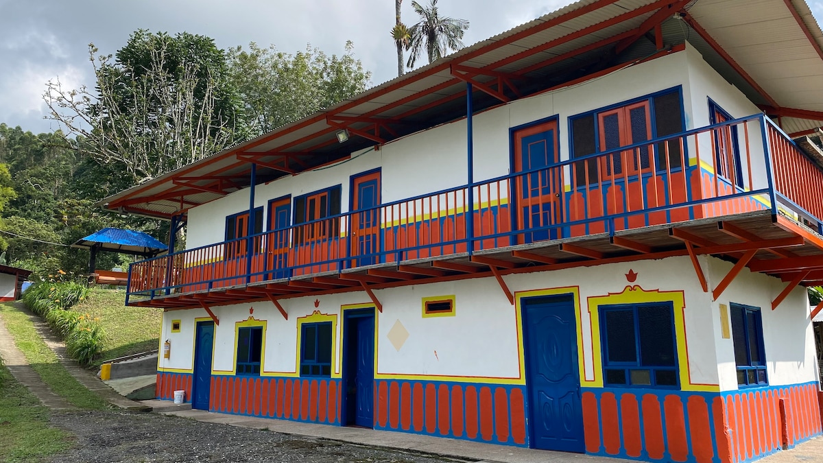 A colorful two-story building is surrounded by greenery, featuring vibrant blue and orange accents. The exterior includes multiple doors and windows, with a balcony extending along the upper level. A pathway leads towards the structure, adding to its welcoming presence.