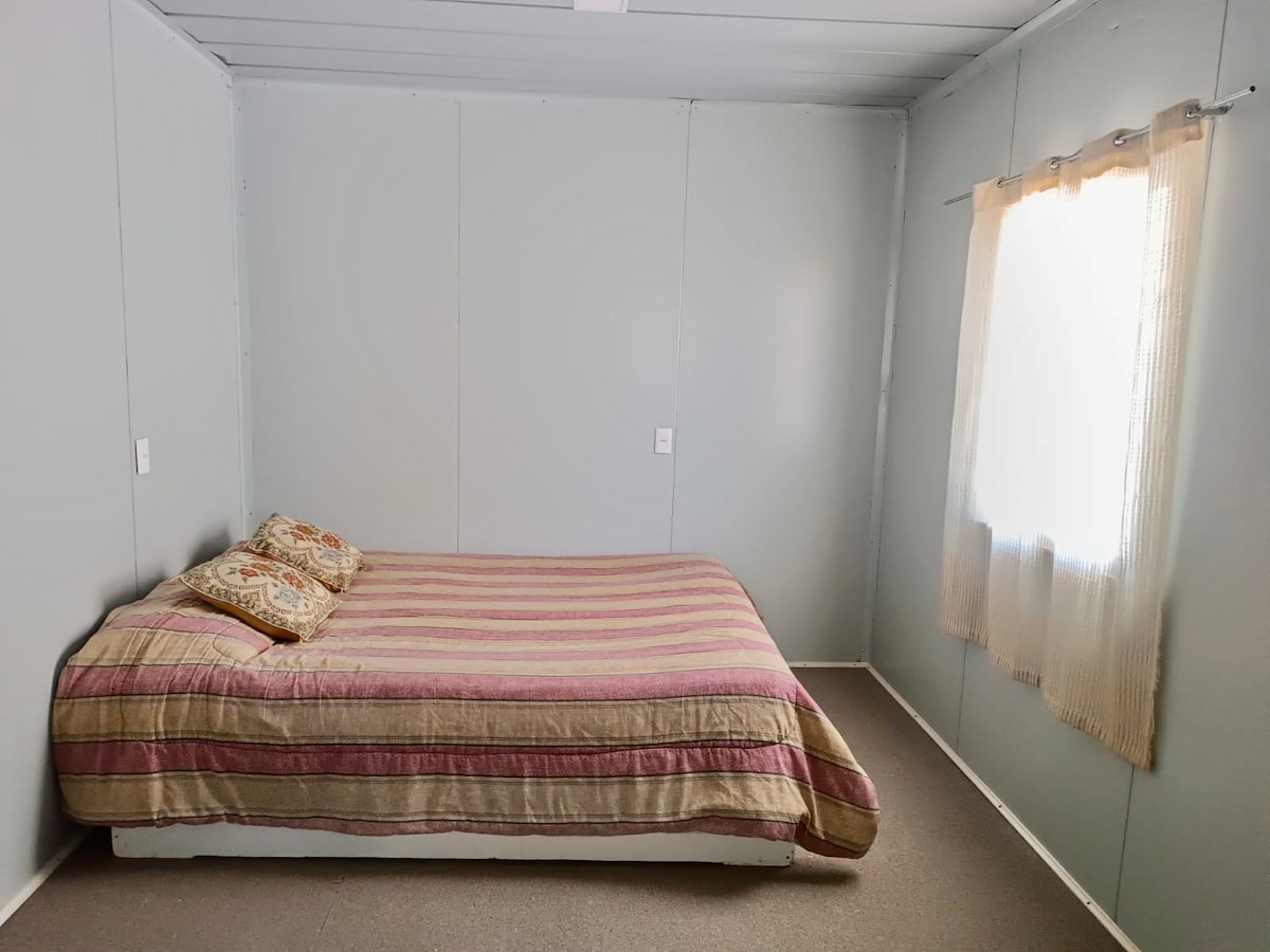 A simple room features a spacious bed with a soft-striped blanket and patterned cushions. Natural light enters through a window adorned with light curtains, illuminating the lightly colored walls and creating a calming environment.