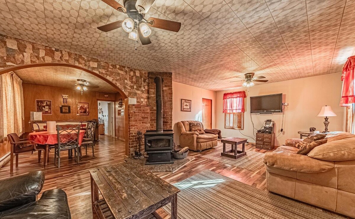 The spacious living area features a wood-burning stove surrounded by comfortable seating, including a sectional sofa and armchairs. A rustic coffee table sits in the center, and large windows with red curtains allow for natural light. The adjacent dining area is visible in the background.