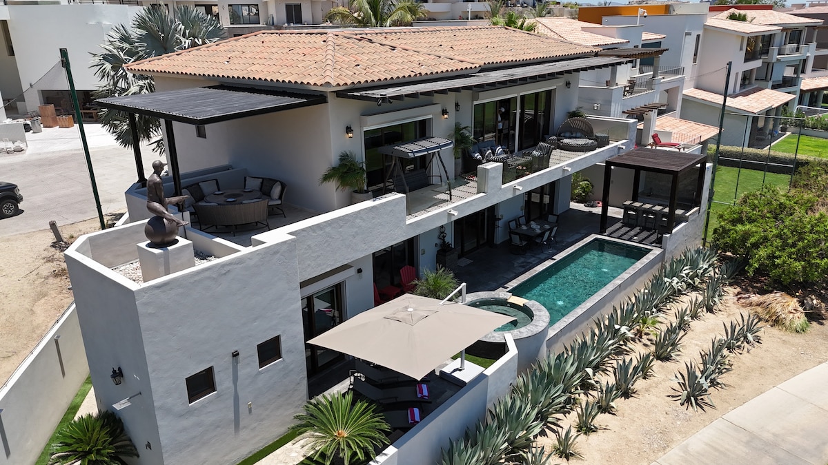 The property features a modern two-story house with a terracotta-tiled roof. A private pool is visible in the backyard, surrounded by lounge chairs and shaded seating areas. The outdoor spaces are complemented by lush landscaping and a view of neighboring structures.