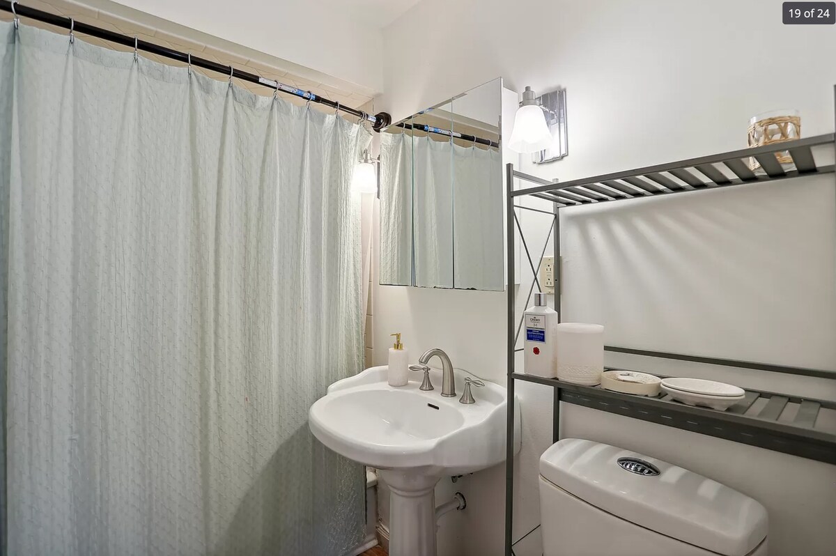 A clean and functional bathroom is shown, featuring a white sink with a mirror above it. A pale blue shower curtain separates the shower area, while a metal shelving unit holds toiletries and towels. Additional amenities are neatly arranged on the shelf.