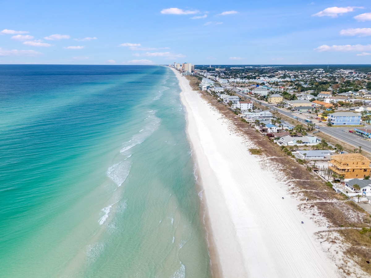 Aerial view captures the expansive shoreline of Panama City Beach, showcasing soft white sand and gentle waves meeting the turquoise waters. The distant skyline features various buildings along the beach, while the coastline extends into the horizon, highlighting the area’s inviting atmosphere.