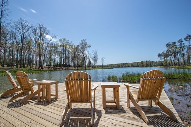 A wooden dock extends over calm waters, furnished with four Adirondack chairs and a small table. Surrounding trees create a natural backdrop, with hints of reflection visible in the water, under a clear blue sky.