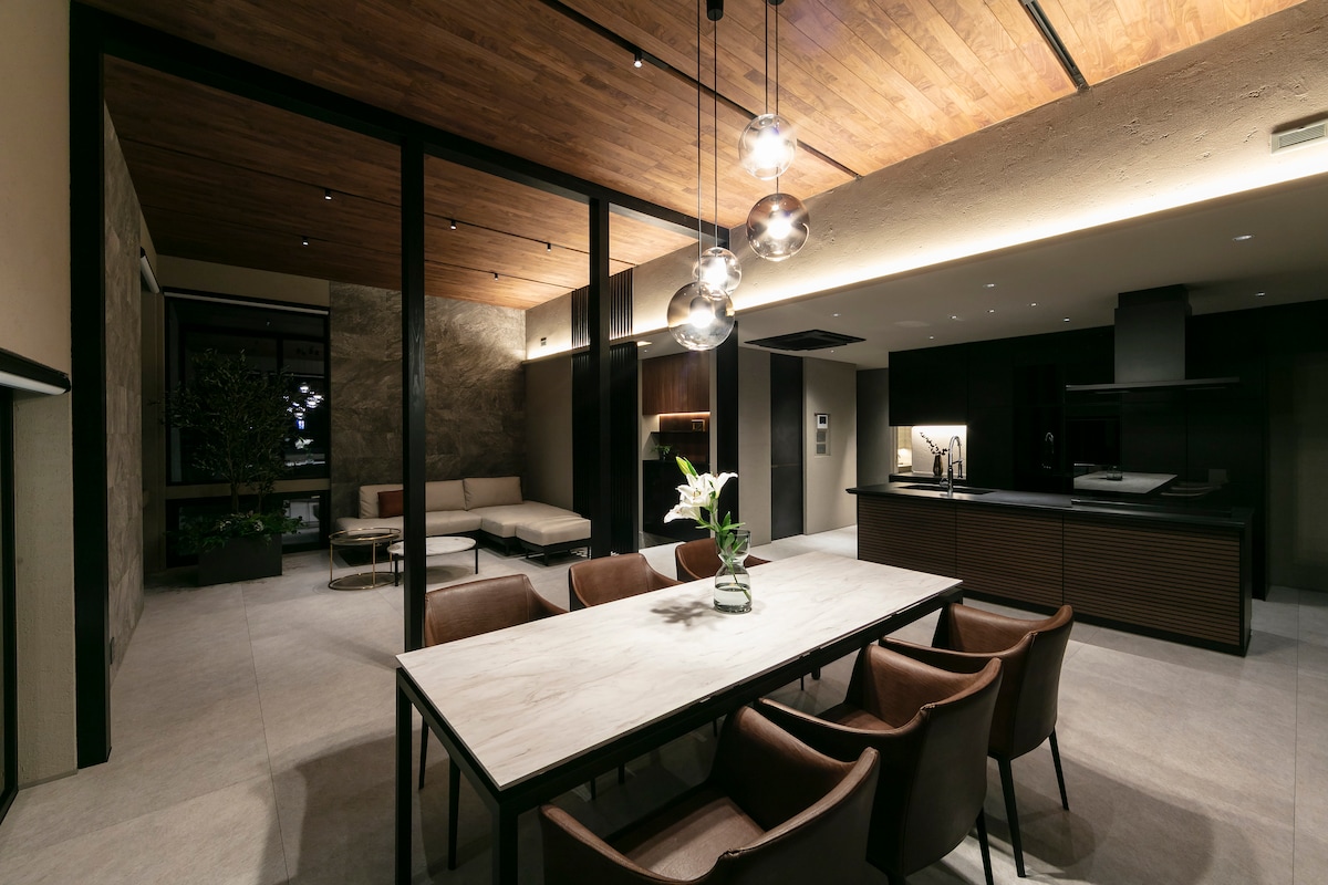 An open-concept dining area and kitchen is highlighted by a spacious marble dining table surrounded by elegant chairs. Soft lighting from modern fixtures complements the natural elements of wood and stone, creating a balanced ambiance. A comfortable seating area is visible in the background.