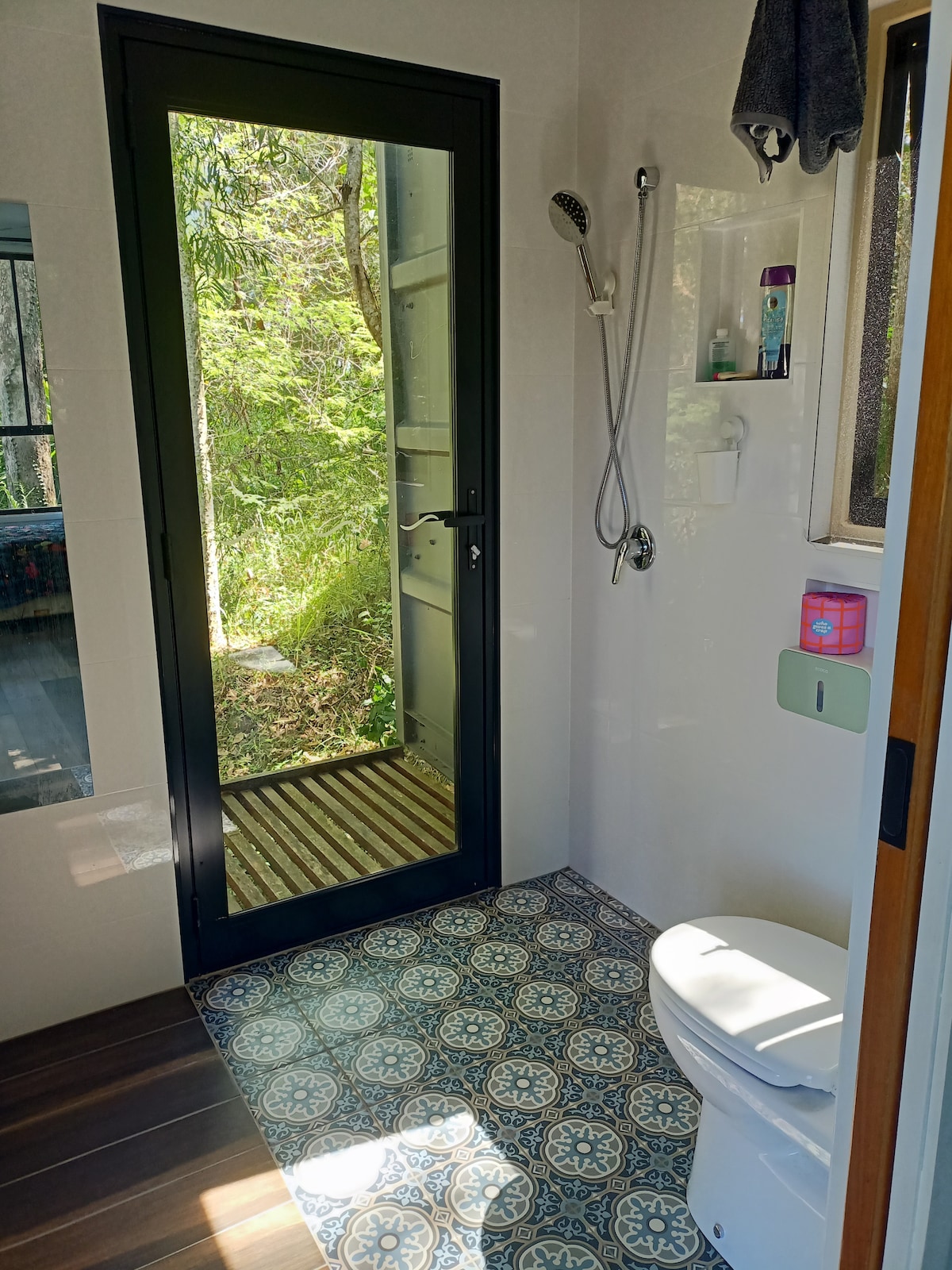 A modern bathroom is showcased, featuring a clean toilet and a shower area with a glass door. Vibrant patterned tiles cover the floor. Natural light filters through the door, illuminating greenery visible from outside, enhancing the serene atmosphere.