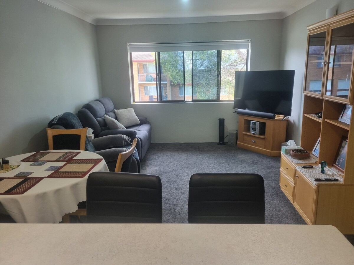 A spacious living area features a comfortable gray sofa and a large television against a neutral wall. A round dining table with chairs is positioned nearby. Natural light enters through a large window, illuminating the carpeted floor and surrounding furnishings.