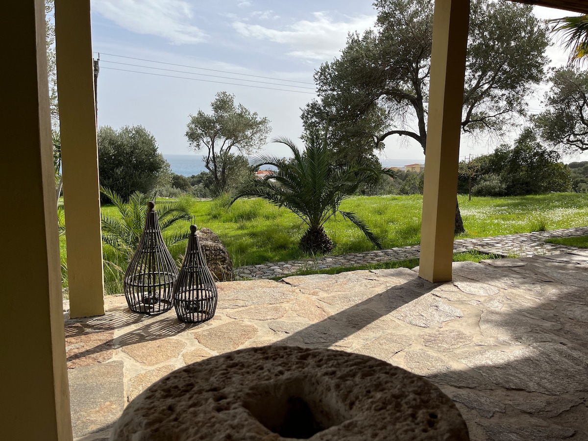An outdoor area features a stone patio overlooking a lush green landscape. Two woven baskets are positioned beside a natural stone element. The distant view includes the Aegean Sea and surrounding trees, enhancing the serene setting.