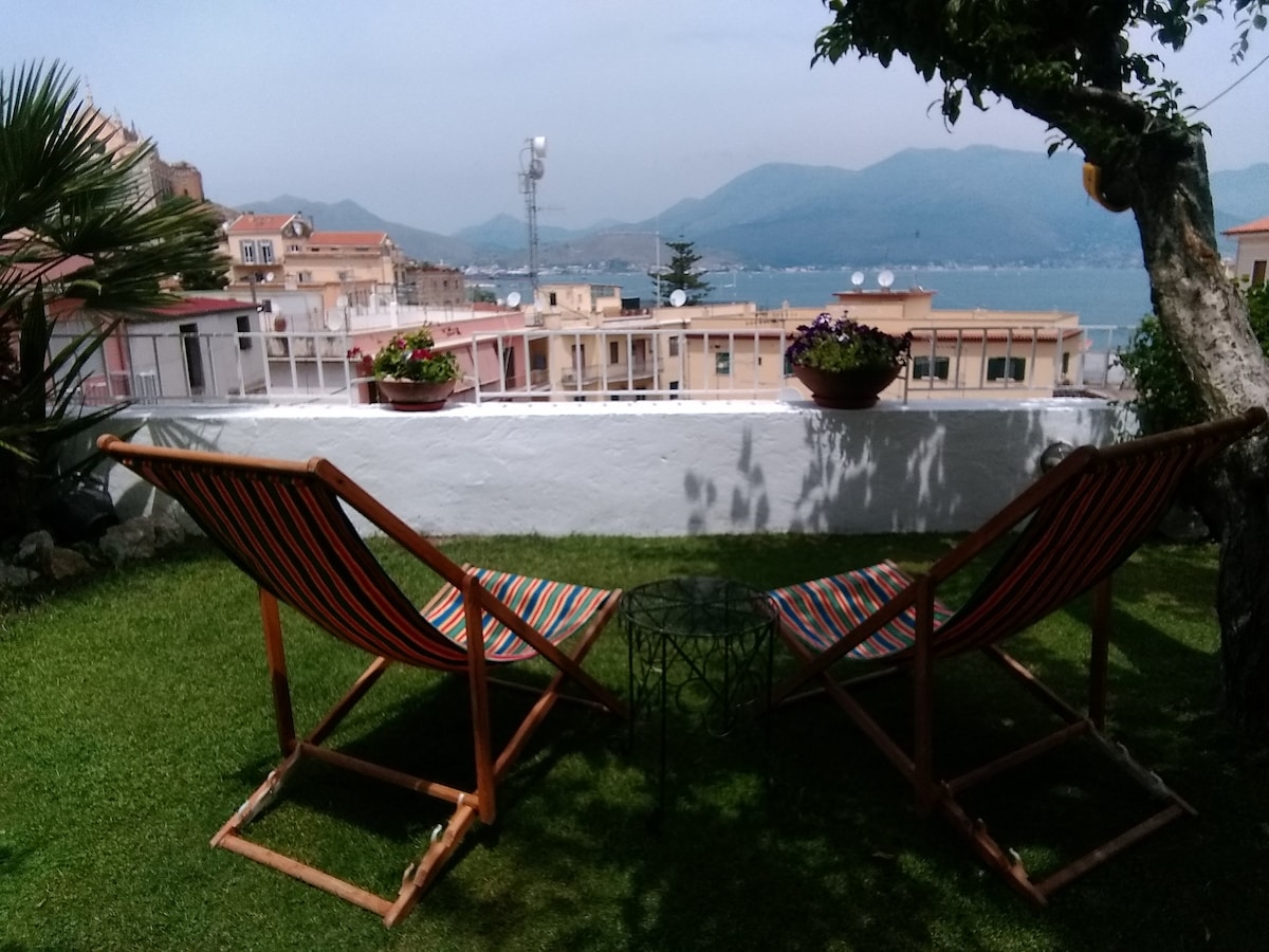 Two striped lounge chairs are positioned on a grassy area, facing a stunning view of the sea and mountains. A small round table sits between the chairs, complementing the setting. Flower pots are placed along the white wall, enhancing the outdoor space.