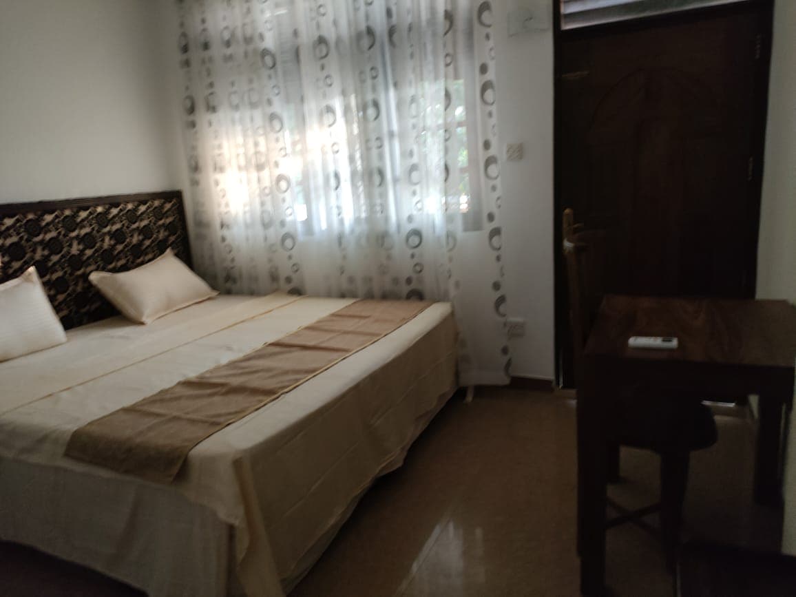 A bright bedroom features a spacious bed with light-colored linens and decorative pillows. Sheer curtains allow natural light to filter in over the neutral walls. A small wooden table with a stool is positioned near the entrance, providing a simple work area.