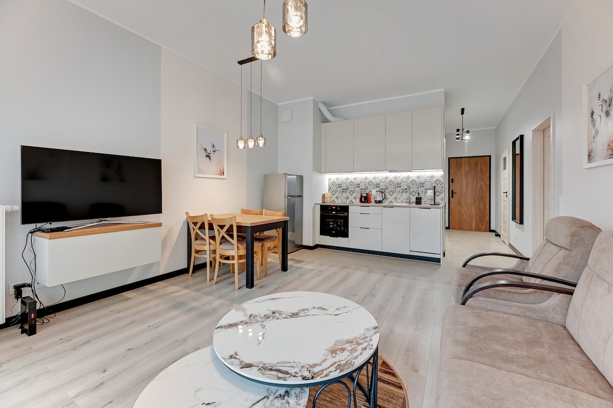 A modern living area is presented with an open layout. A large flat-screen TV is mounted on the wall opposite a comfortable sofa and armchairs. A dining table with wooden chairs is positioned near the kitchen, which features sleek cabinetry and modern appliances.