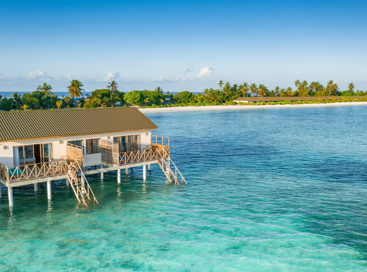 A water villa is situated above clear turquoise waters, supported by stilts. Lush tropical vegetation can be seen along the shoreline, with a sandy beach in the background. The villa features a sloped roof, large windows, and a wooden deck extending over the water.