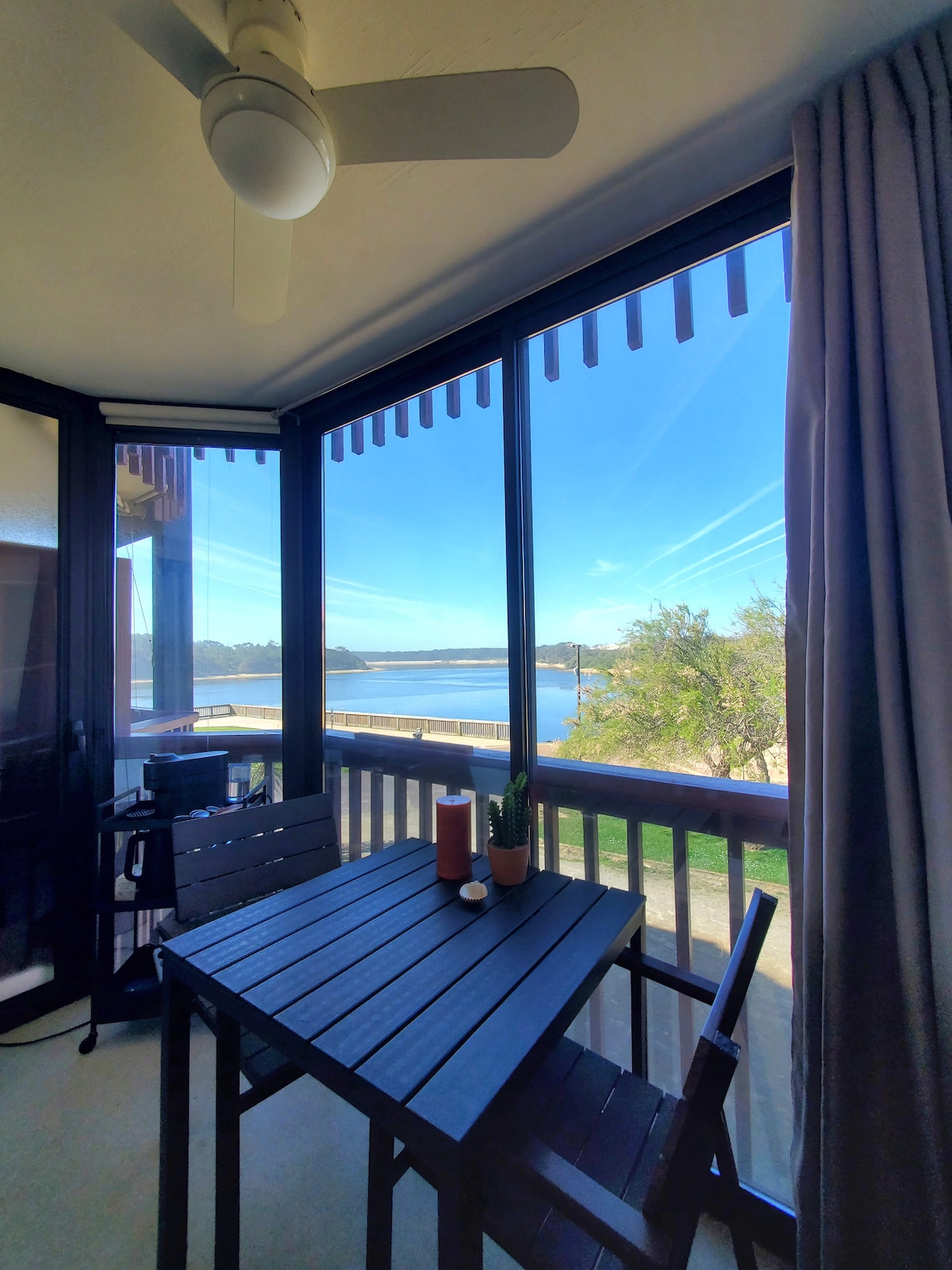 A serene enclosed veranda features a wooden table with two chairs, accompanied by decorative plants. Large windows provide an unobstructed view of the lake and surrounding landscape, allowing ample natural light to fill the space.