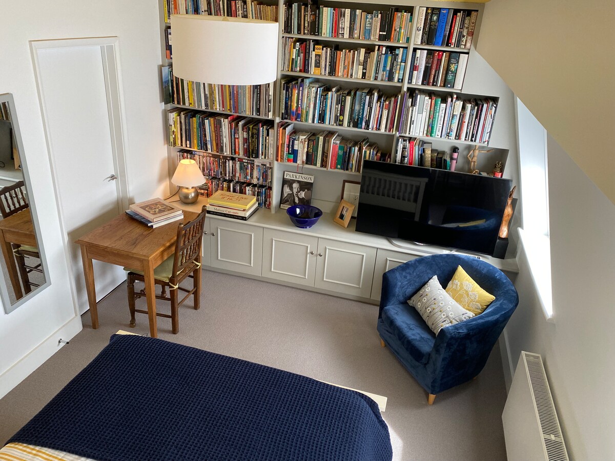 A well-organized bedroom features a wall of bookshelves filled with various books. A small wooden table with a chair is positioned in the corner. A spacious bed is visible, and a blue armchair with a decorative pillow is nearby. A TV rests on a cabinet across from the bed.