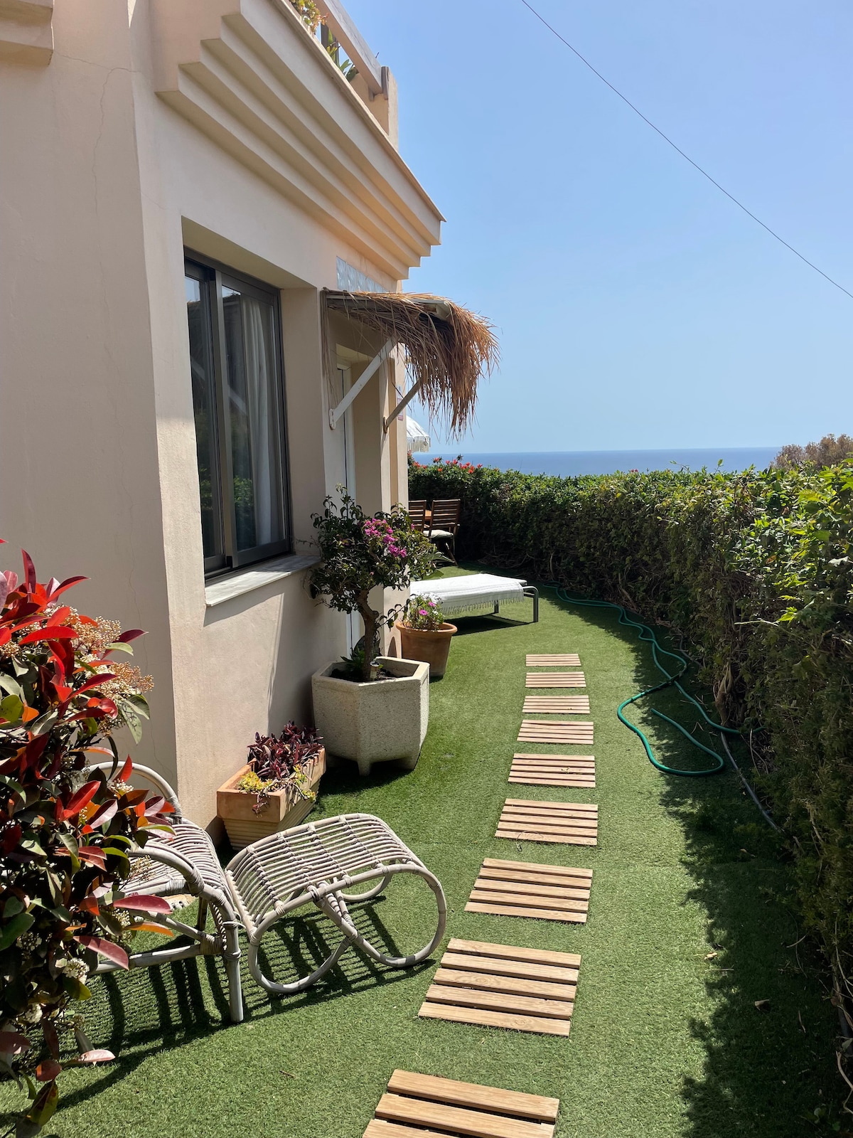 A neatly arranged outdoor space is visible, featuring a pathway of wooden planks leading to a sunbed. Lush greenery surrounds the area, with flowering plants in pots adding color. A gentle view of the sea can be seen in the background under a clear blue sky.
