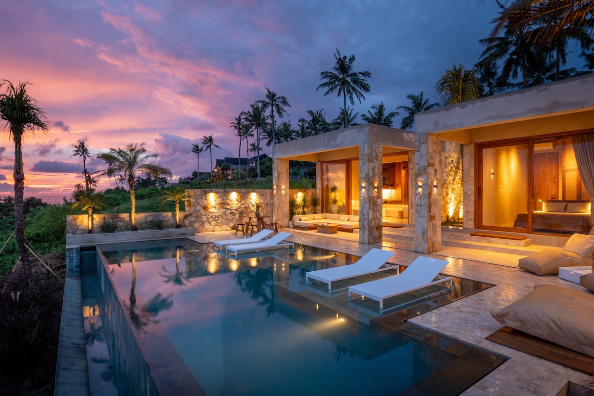 An elegantly designed villa is set against a vibrant sunset sky, with a private pool reflecting warm colors. Lounge chairs are positioned near the water, while palm trees line the property, enhancing the tranquil atmosphere of the outdoor space.
