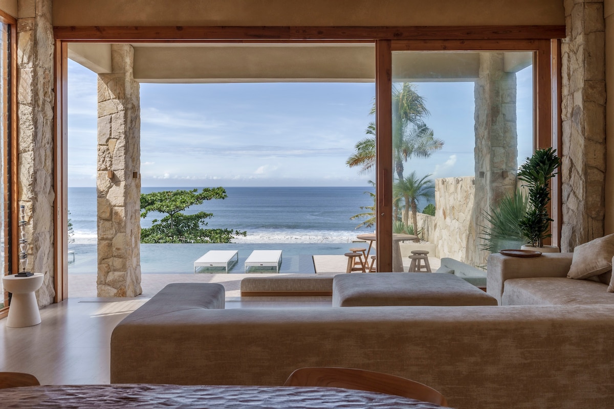 A spacious living area opens up to breathtaking ocean views through large glass doors. Natural stone walls complement the tranquil ambiance, while comfortable seating invites relaxation. The outdoor pool and lounge chairs are visible, framed by vibrant greenery and soft coastal scenery.