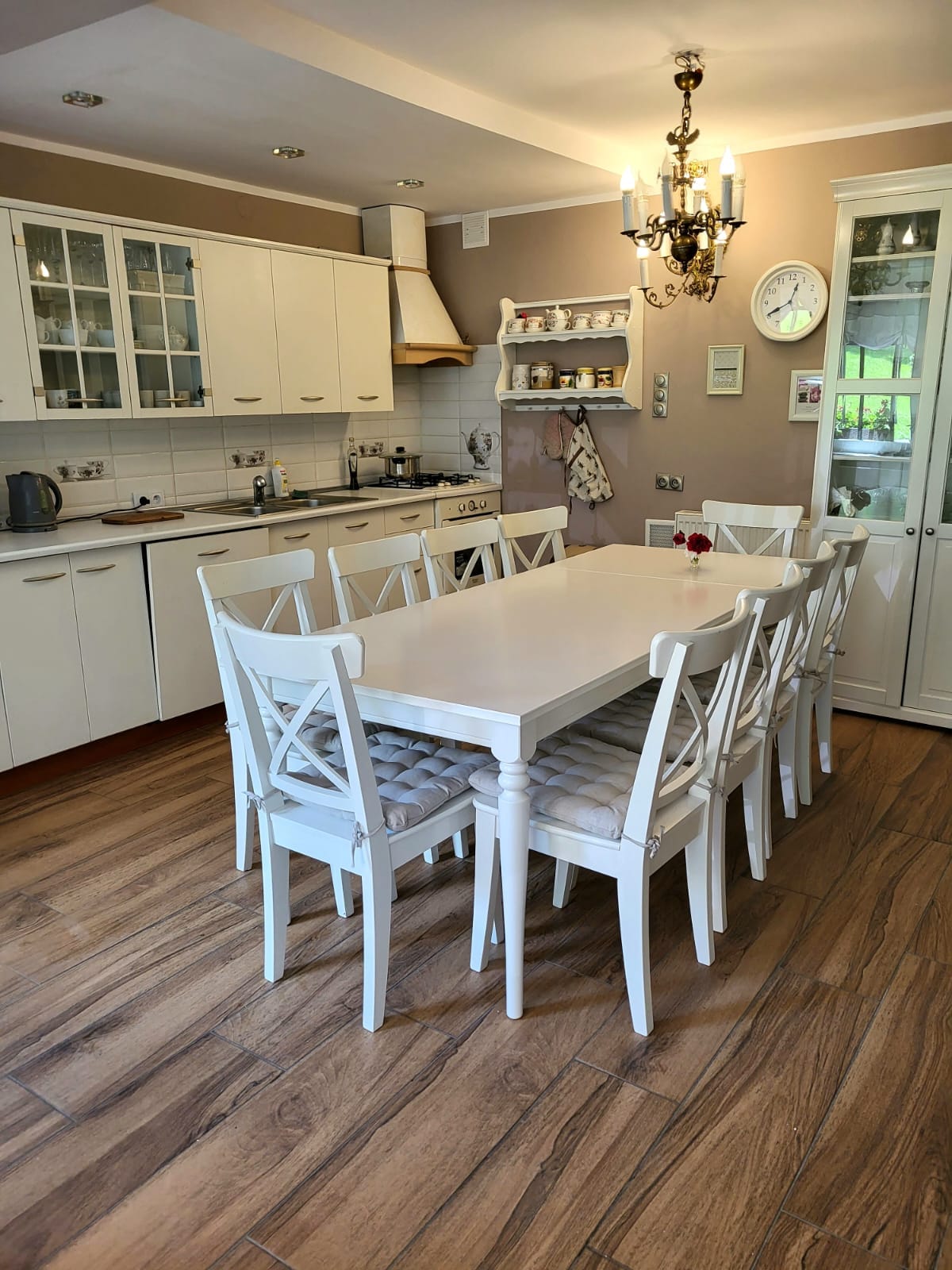 A modern kitchen is showcased, featuring white cabinetry and a spacious dining table surrounded by six matching chairs. A chandelier hangs overhead, illuminating the room. Cabinets with glass fronts display neatly arranged dishware, while countertops hold essential kitchen appliances.