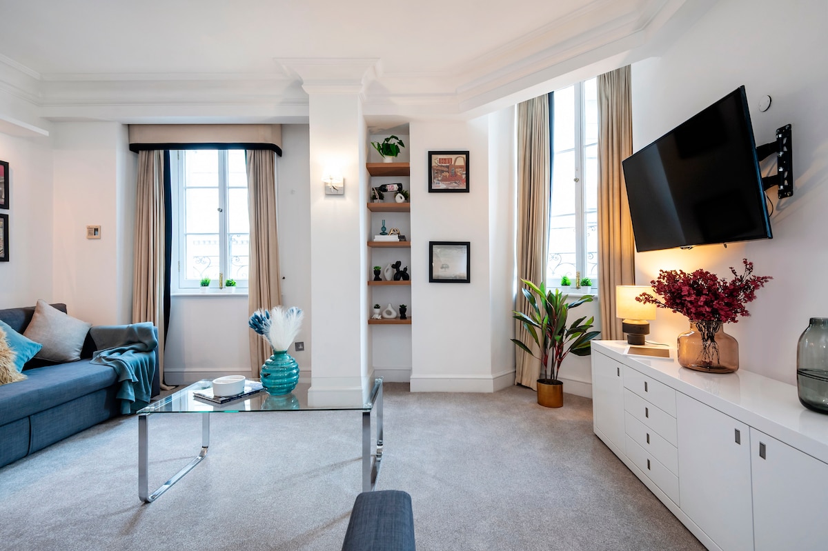 A modern living area is presented with a plush gray sofa and a glass coffee table. Light flows through two tall windows dressed with drapes. Decorative shelves display framed art and plants, while a flat-screen TV is mounted on the wall beside a stylish console.