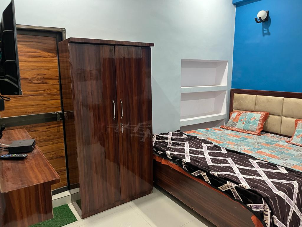 A cozy bedroom features a comfortable double bed with patterned bedding against a vibrant blue wall. A wooden wardrobe stands nearby, and a TV is mounted above a desk with a polished surface, enhancing the room's functionality.