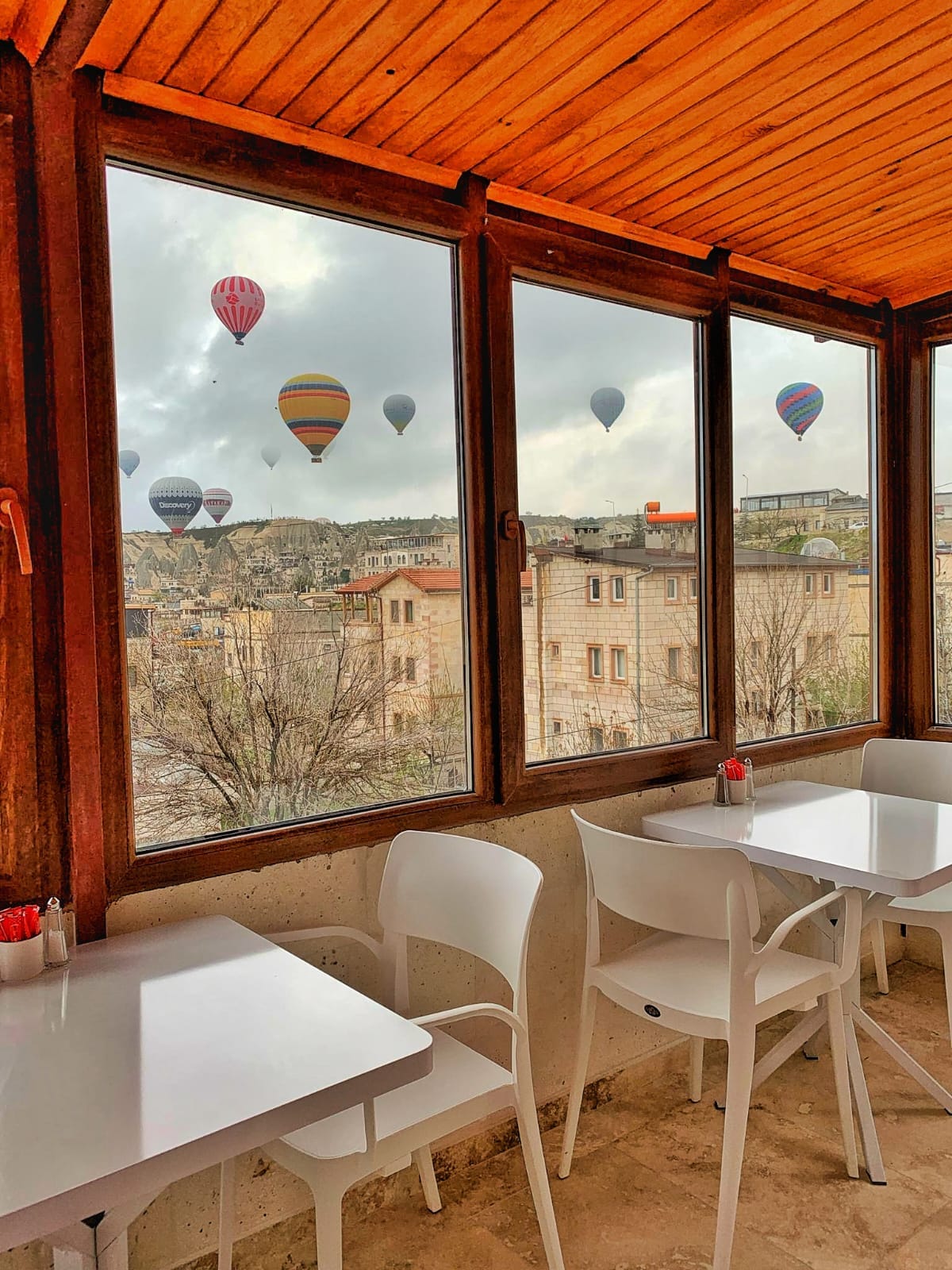 Large windows frame a view of colorful hot air balloons floating in the sky. A bright dining area features three simple white tables and chairs, enhancing the serene setting. The surrounding buildings showcase traditional architectural details, adding charm to the view.
