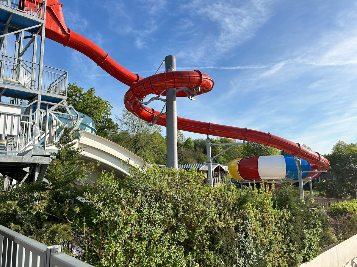 A vibrant water slide structure is depicted, featuring a series of twisting red slides along with multi-coloured slides. Lush green foliage surrounds the area, providing a natural contrast to the bright slides and enhancing the sense of outdoor fun.
