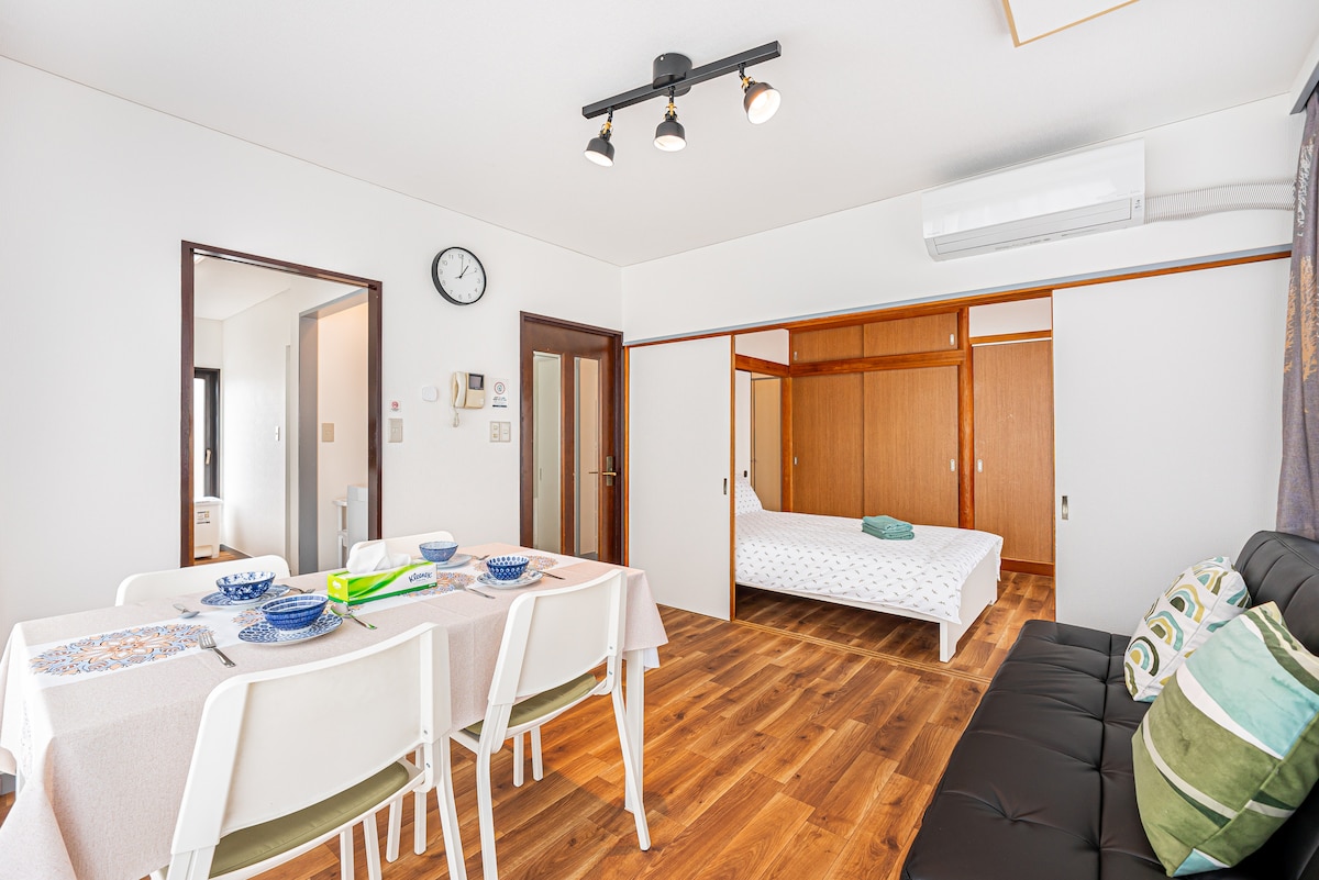 A bright living area is shown with wooden flooring, featuring a dining table set for six. A black sofa adds contrast, while a spacious bed is visible in the adjacent area. Light is provided by modern track lighting and natural light from the windows.
