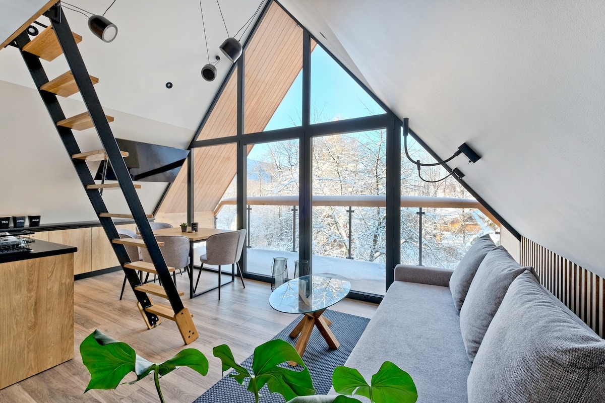 A modern loft space is highlighted by large windows offering a view of snow-covered trees. A light gray sofa sits beside a circular glass coffee table. A dining area with a wooden table and two chairs is visible, along with a stylish staircase leading to the upper level.