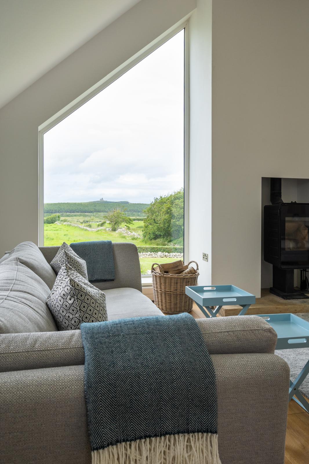 A light-filled living area features large angled windows showcasing lush green landscapes. A grey sofa is adorned with textured cushions, complemented by two small blue tables nearby. A cozy throw drapes over the arm of the sofa, adding warmth to the space.