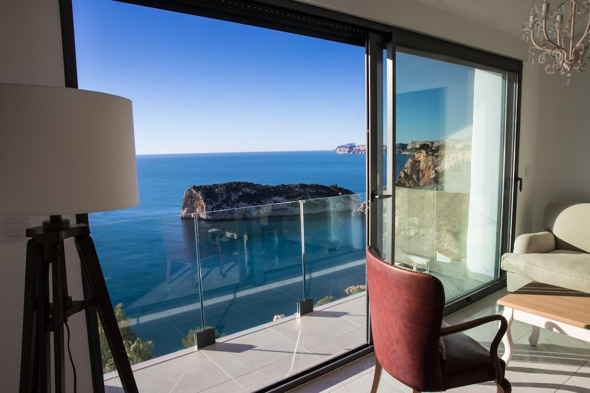 A bright interior space offers panoramic views of the sea through large glass doors. A comfortable armchair and stylish lamp create a warm corner, while the striking coastal scenery invites relaxation and contemplation.