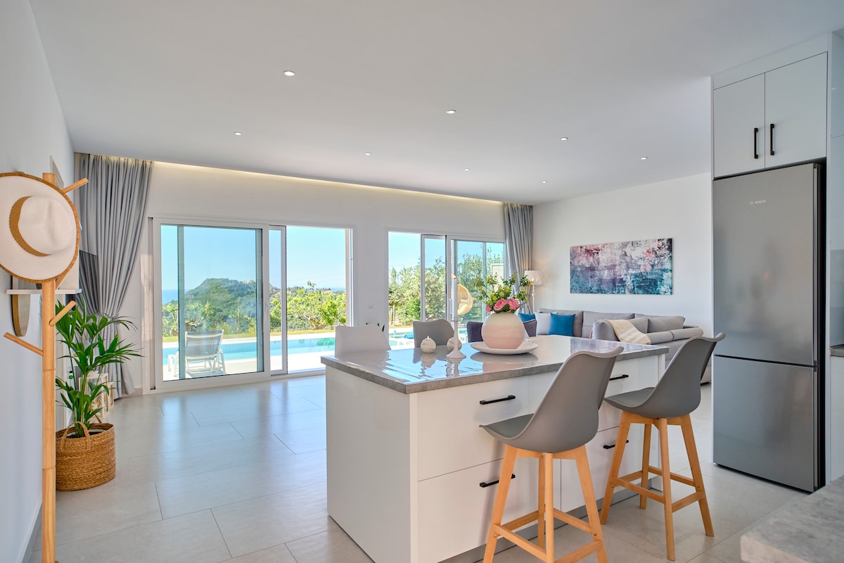 A spacious kitchen and living area are showcased, featuring large glass doors that open to the outdoor pool area. The kitchen island is complemented by modern bar stools, while natural light illuminates the space. A decorative plant adds a touch of greenery.