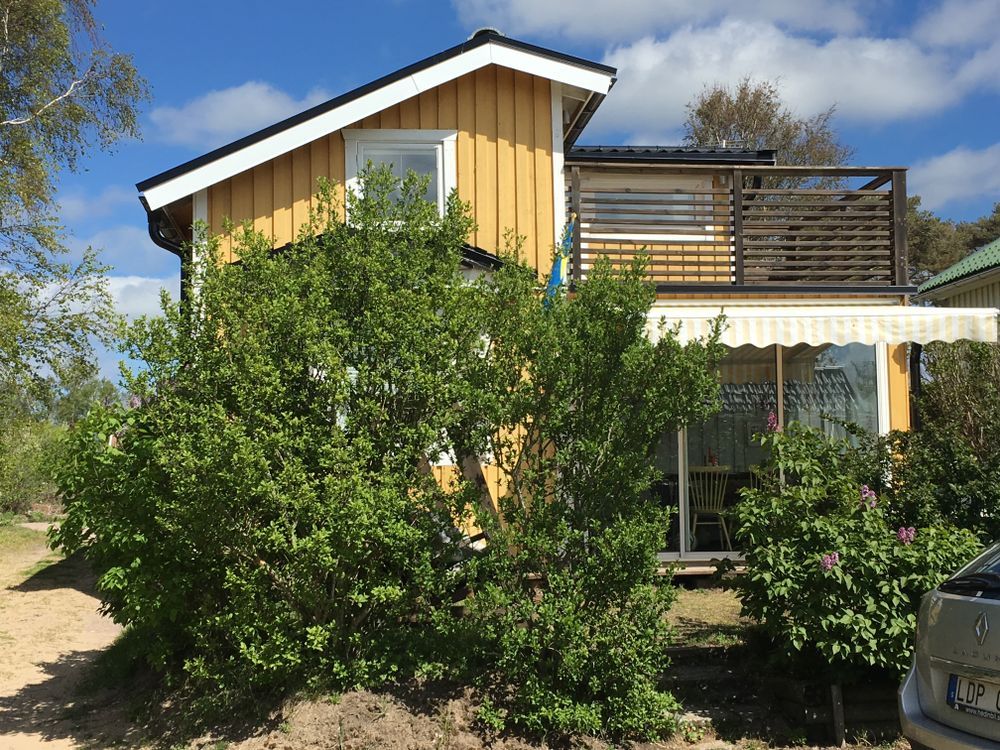 The exterior of a two-story yellow cottage is surrounded by greenery. A covered terrace is visible on the ground floor, providing a shaded seating area. The upper level features a wooden deck with outdoor furniture. A bright blue sky and scattered clouds enhance the cheerful atmosphere.