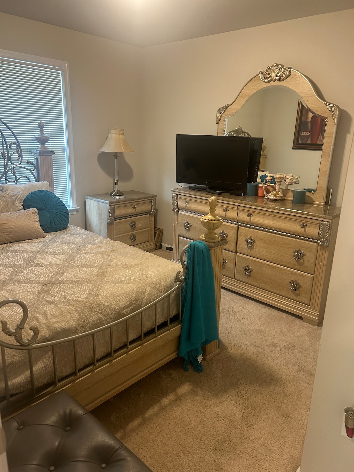 A neatly arranged bedroom features a spacious bed with an ornate metal frame, soft bedding, and decorative pillows. Nearby, a dresser displays a small television and personal decor. A bedside lamp provides additional lighting, while natural light enters through a window with blinds.