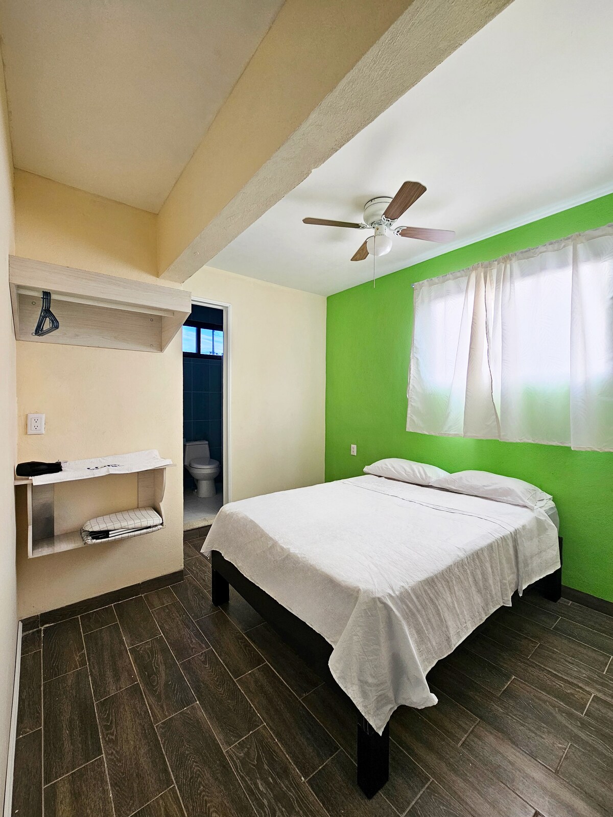 A bright bedroom features a spacious bed covered with a white blanket. A fan hangs from the ceiling, and a small shelf is positioned near the bed. A window allows natural light to fill the room, complemented by a vibrant green wall. A bathroom is visible through an open door.