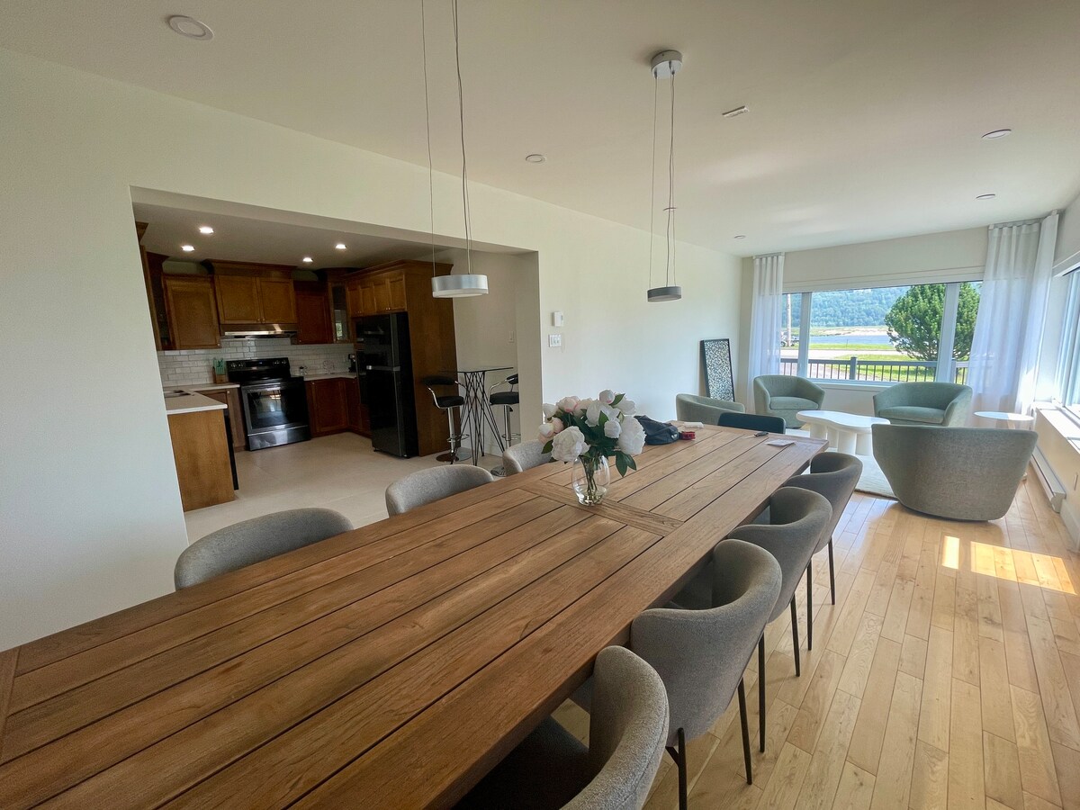 A spacious dining area showcases a long wooden table surrounded by ten upholstered chairs. In the background, an open concept kitchen features modern appliances. Large windows allow natural light to fill the space, providing views of the outdoors.