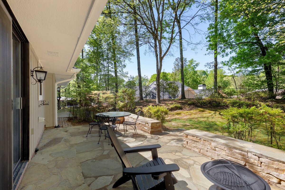A stone patio is presented with outdoor seating, including black chairs and a table. Lush greenery surrounds the area, offering a serene view of the landscaped yard. Sunlight filters through the trees, creating a peaceful outdoor space for relaxation.