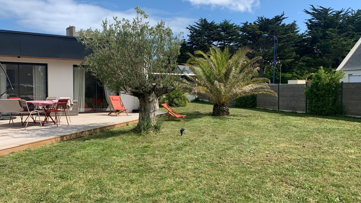 The outdoor space features a well-maintained grassy garden, surrounded by palm trees and an olive tree. Red lounge chairs are positioned on a wooden terrace, which provides an area for outdoor dining with a table and chairs, creating a relaxing environment.