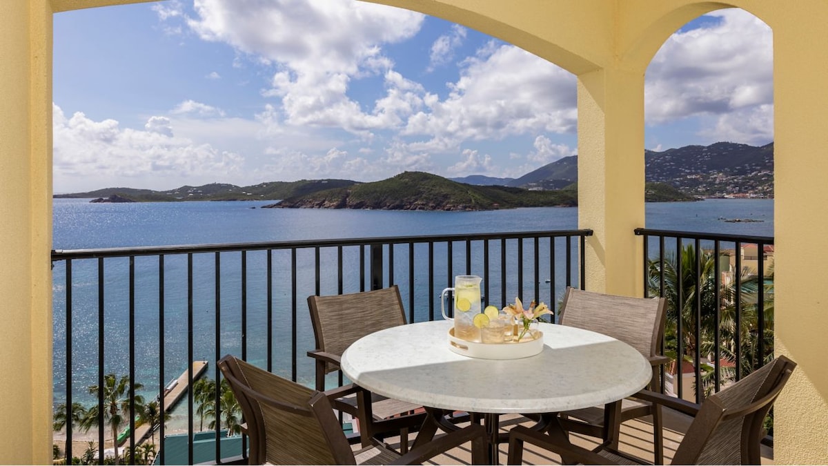 A private balcony offers a round table set with four chairs, overlooking the tranquil blue waters of Pacquereau Bay. Scenic views of distant islands are framed by a curved archway, with partial clouds visible in the expansive sky.