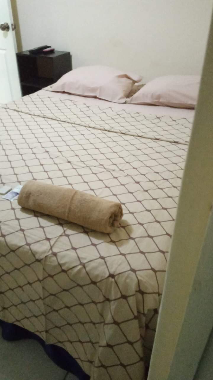 A cozy sleeping area features a spacious bed with a neutral-toned blanket and two soft pillows. A rolled towel is placed neatly on the bed, adding a touch of comfort. A small dark wood cabinet is visible nearby.