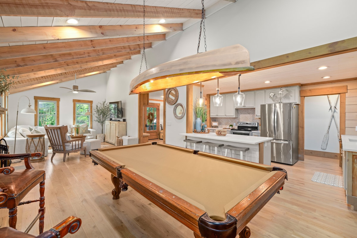 An inviting game room features a classic pool table at its center, illuminated by a unique light fixture resembling a canoe. Light wood beams and ample natural light create a bright atmosphere, while a modern kitchen and seating area are visible in the background.