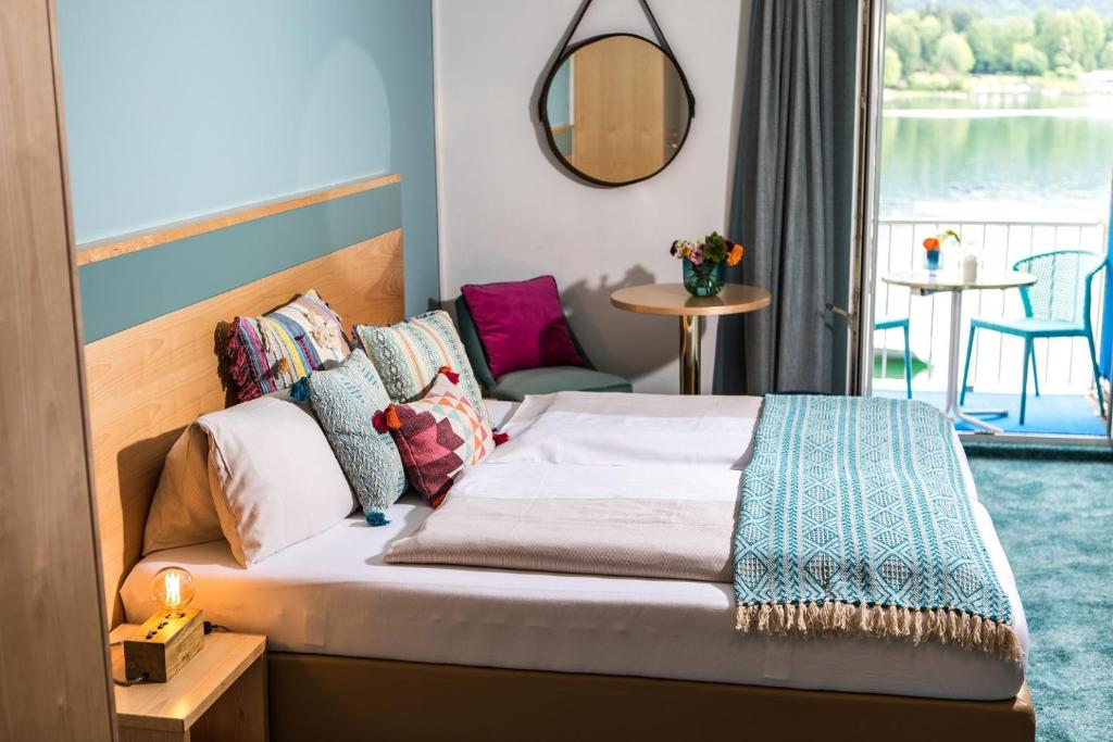 A bright sleeping area features a spacious bed adorned with decorative pillows and a textured throw. A round mirror reflects natural light, while a small table and two chairs overlook the serene lake through large windows.