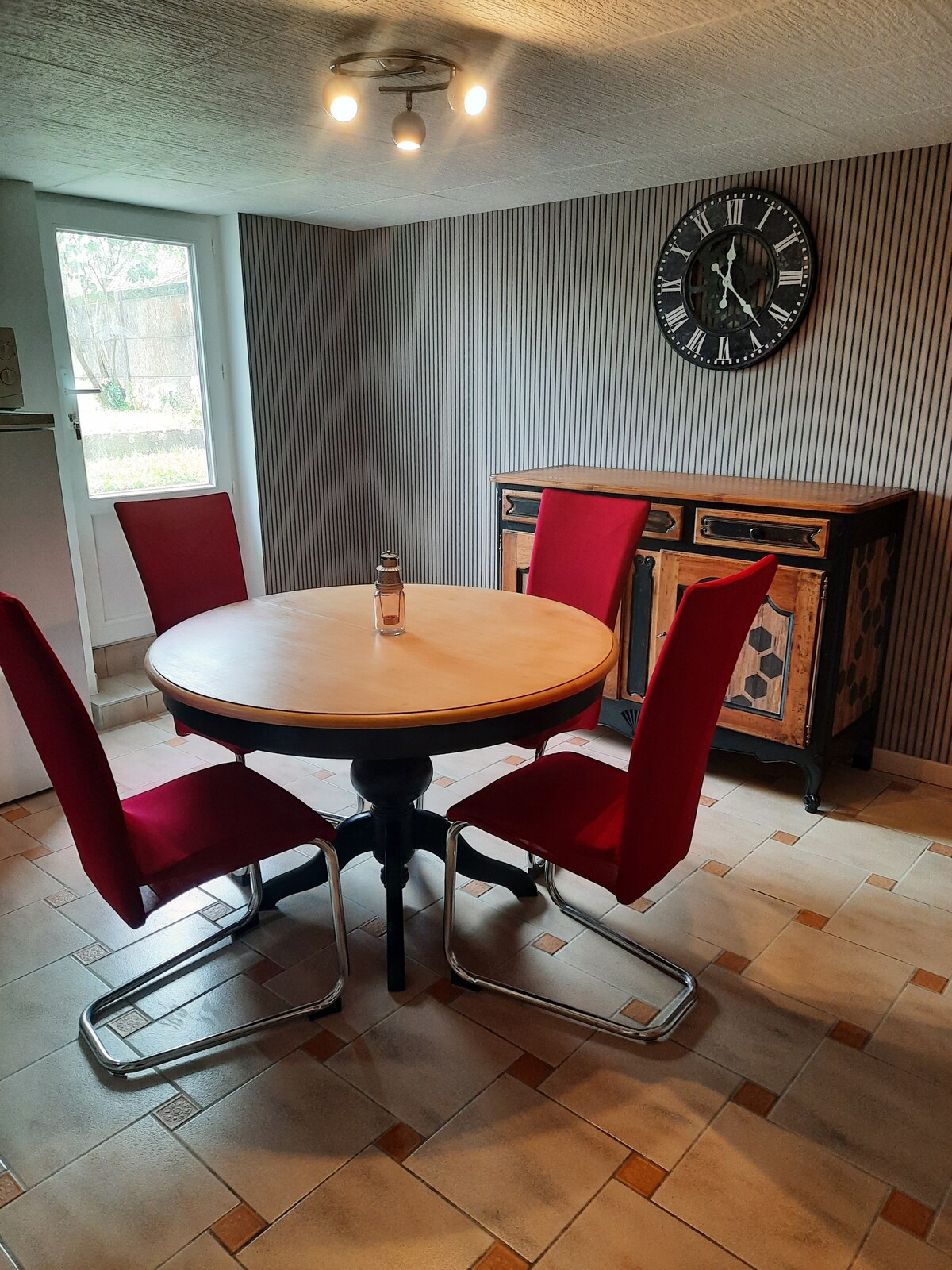 A dining area features a round wooden table surrounded by four red chairs. A side cabinet with decorative elements is positioned against the wall, while a large clock is mounted above it. Natural light enters through a nearby window, illuminating the space.