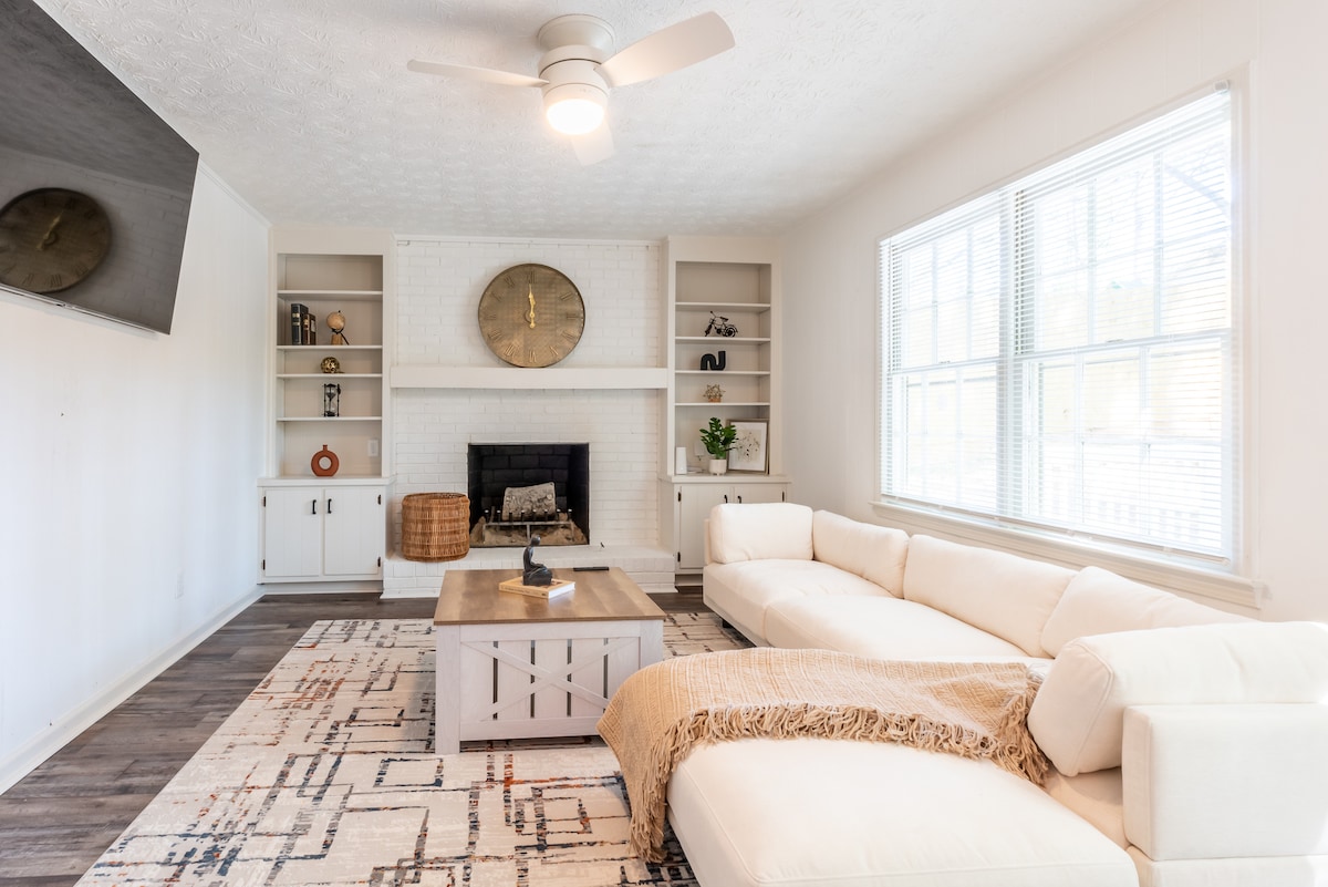 A light-filled living room features a large L-shaped sectional sofa arranged around a coffee table. A modern fan hangs from the ceiling above. The white brick fireplace serves as a focal point, flanked by built-in shelves displaying decorative items.