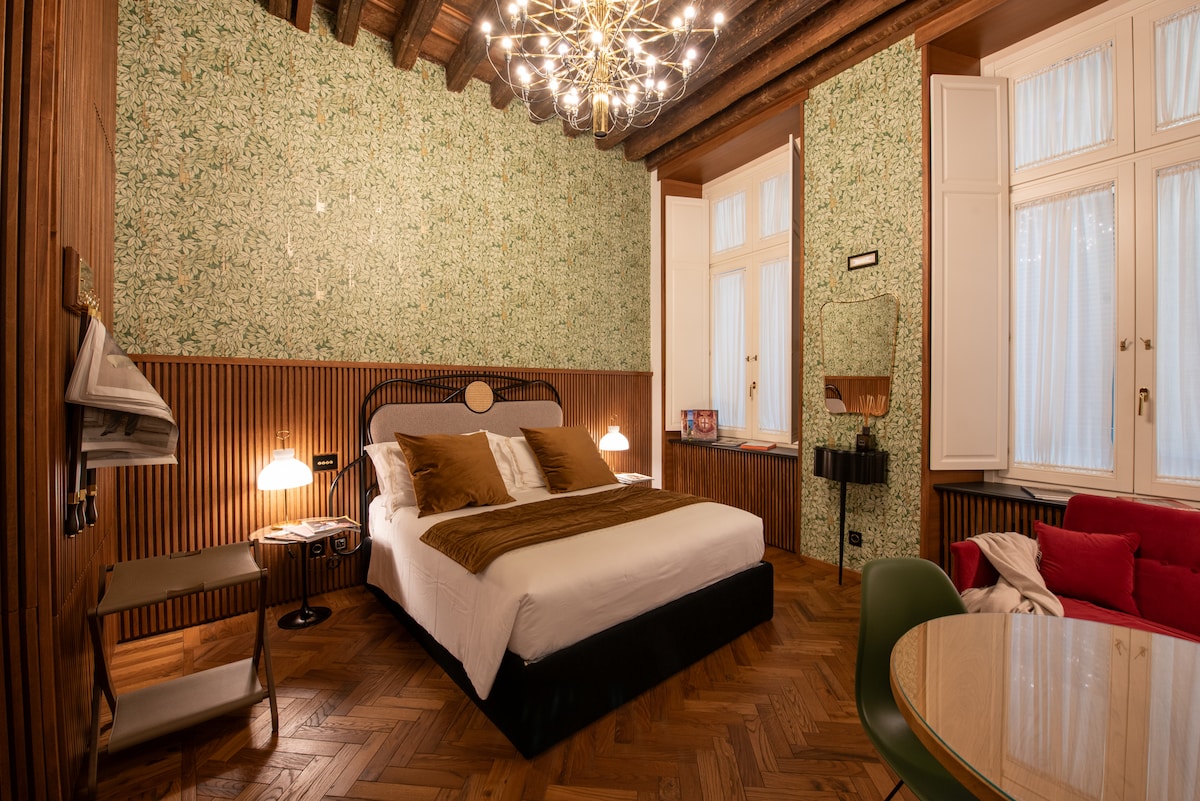 A well-appointed bedroom features a spacious bed with neutral-toned linens, flanked by stylish bedside lamps. The walls are adorned with floral wallpaper, while wood paneling adds warmth. A cozy seating area includes a vibrant red sofa, and natural light filters through multiple windows.