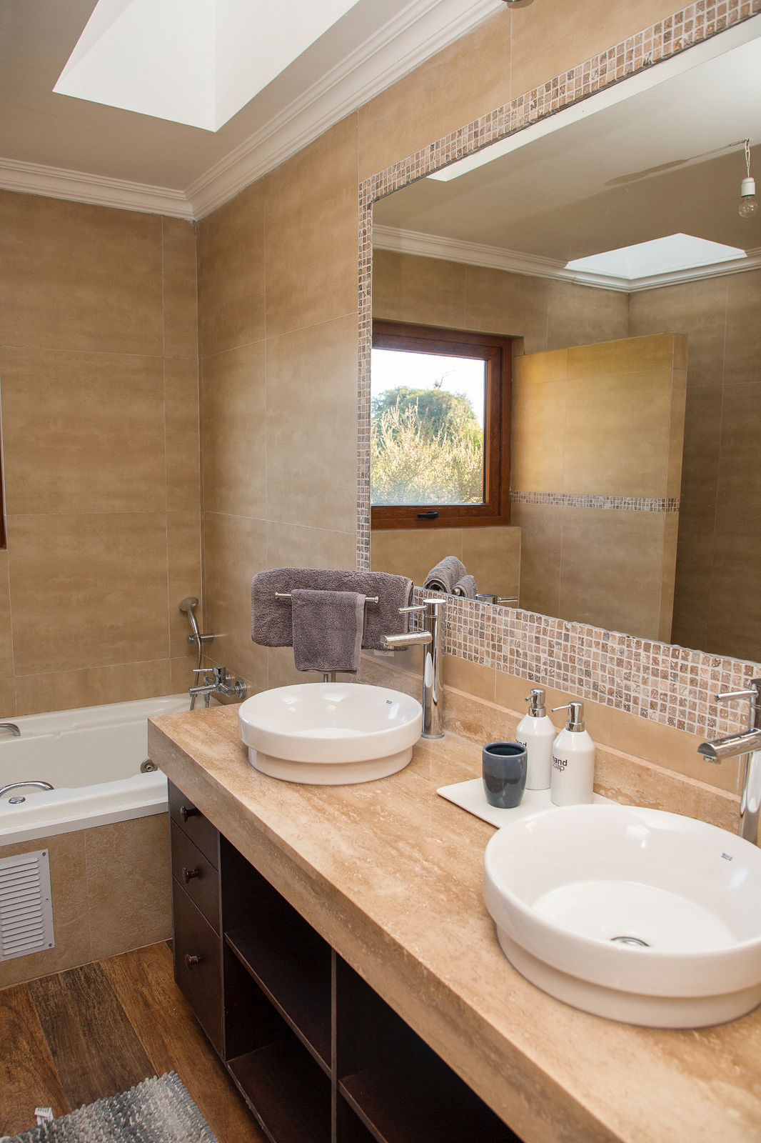 A well-appointed bathroom features dual round sinks positioned on a natural stone countertop. A large mirror reflects the soft light from a skylight above. A soothing color palette is complemented by plush towels, and a soaking tub is visible against an adjoining wall.