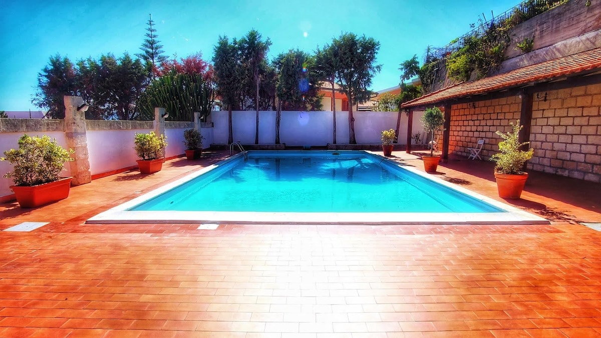 A serene swimming pool is framed by a sunlit patio, featuring terracotta tiles and potted plants. Lush greenery is visible in the background, providing a calming environment. The pool's inviting turquoise water reflects the clear sky overhead, enhancing the outdoor space's inviting feel.